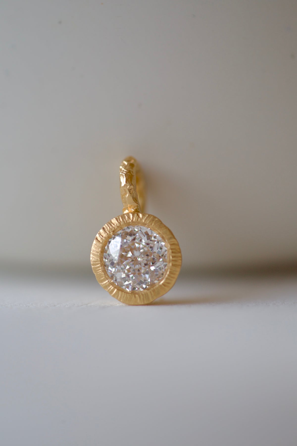 Glypto Diamond Pendant, hammered gold bezel and carved bail with a round diamond, on a cream surface