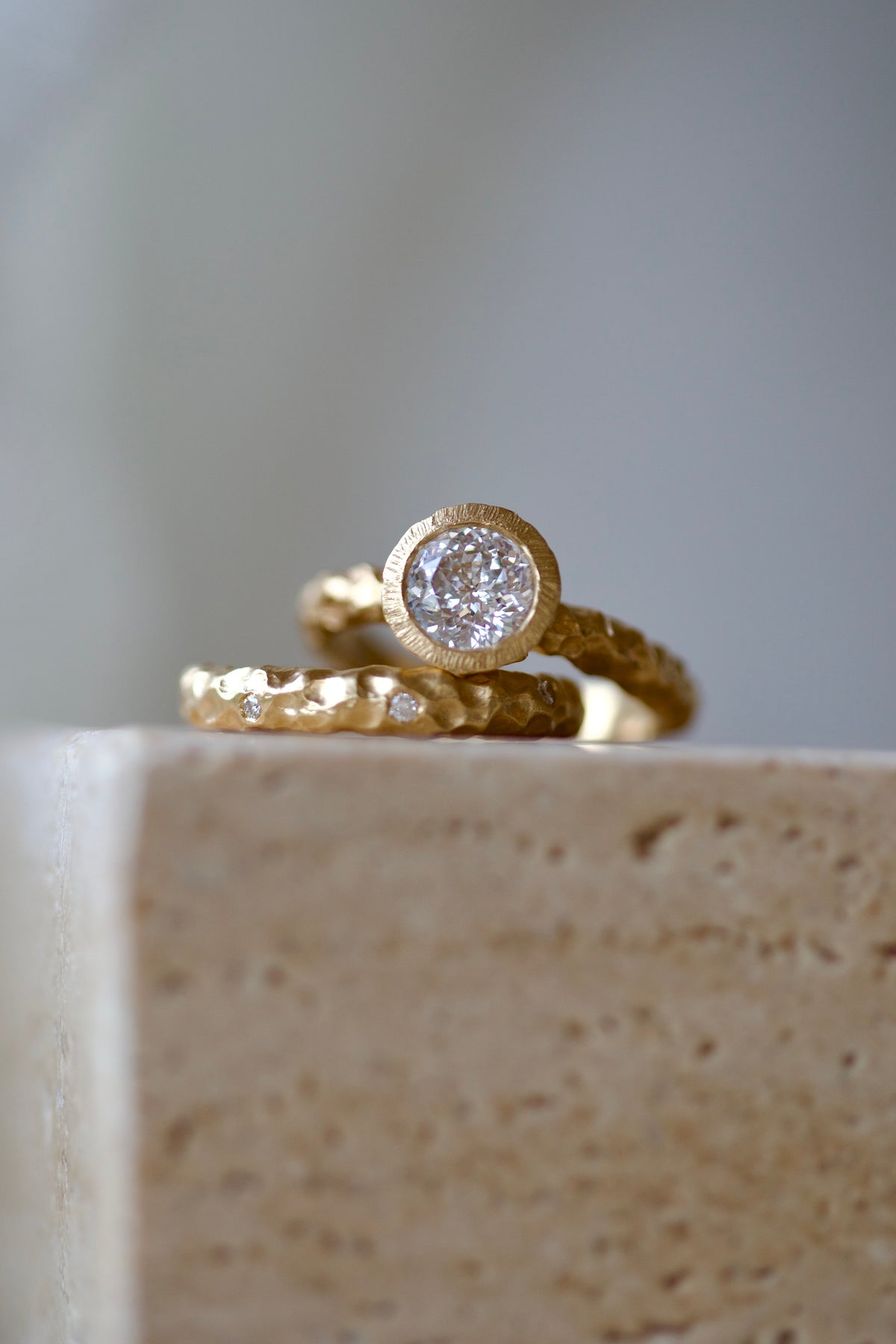 Glypto Solitaire Ring stacked with a hammered gold band set with small diamonds, on a travertine block